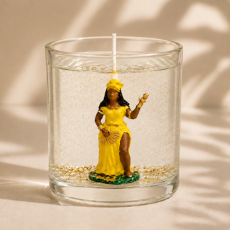 Vela artesanal em gel transparente com imagem de Oxum vestida em amarelo e dourado, posicionada no centro de um recipiente de vidro cilíndrico. O gel contém delicados detalhes dourados suspensos e a composição é fotografada em um ambiente suave e elegante, com iluminação natural e sombras orgânicas que reforçam a sensação de prosperidade, harmonia e sofisticação.