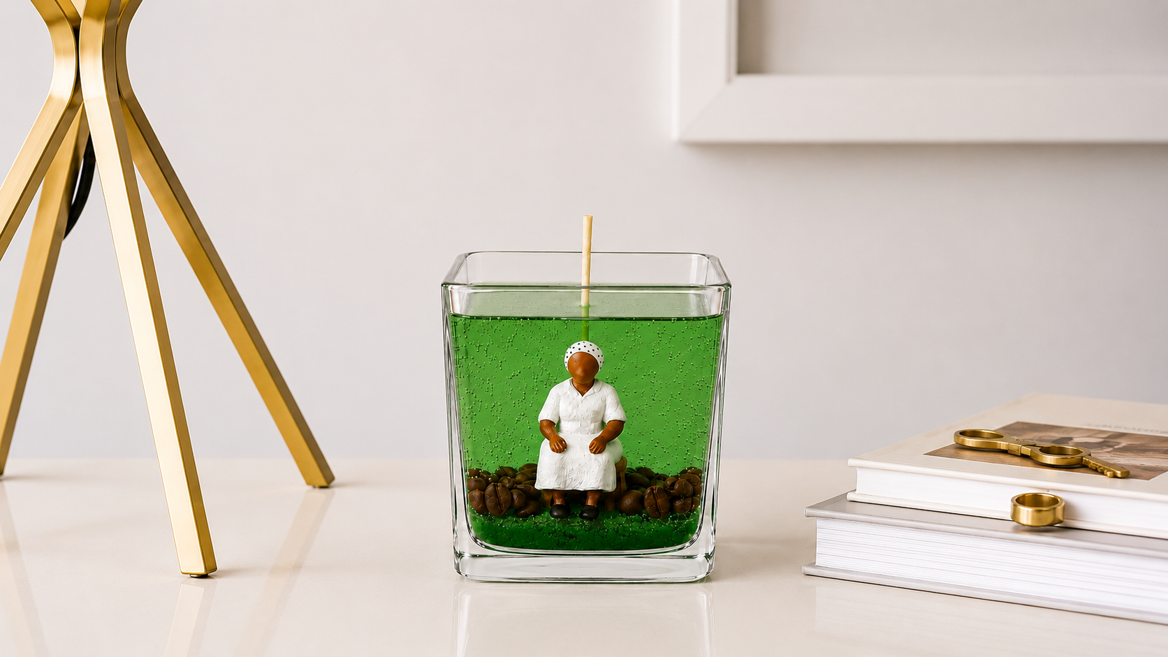 Vela em Gel Preta Velha artesanal em vidro quadrado com parafina em gel verde translúcida, imagem de Preta Velha sentada e grãos de café decorativos. A peça está posicionada sobre uma mesa clara em ambiente sofisticado, com livros e elementos decorativos dourados ao fundo.