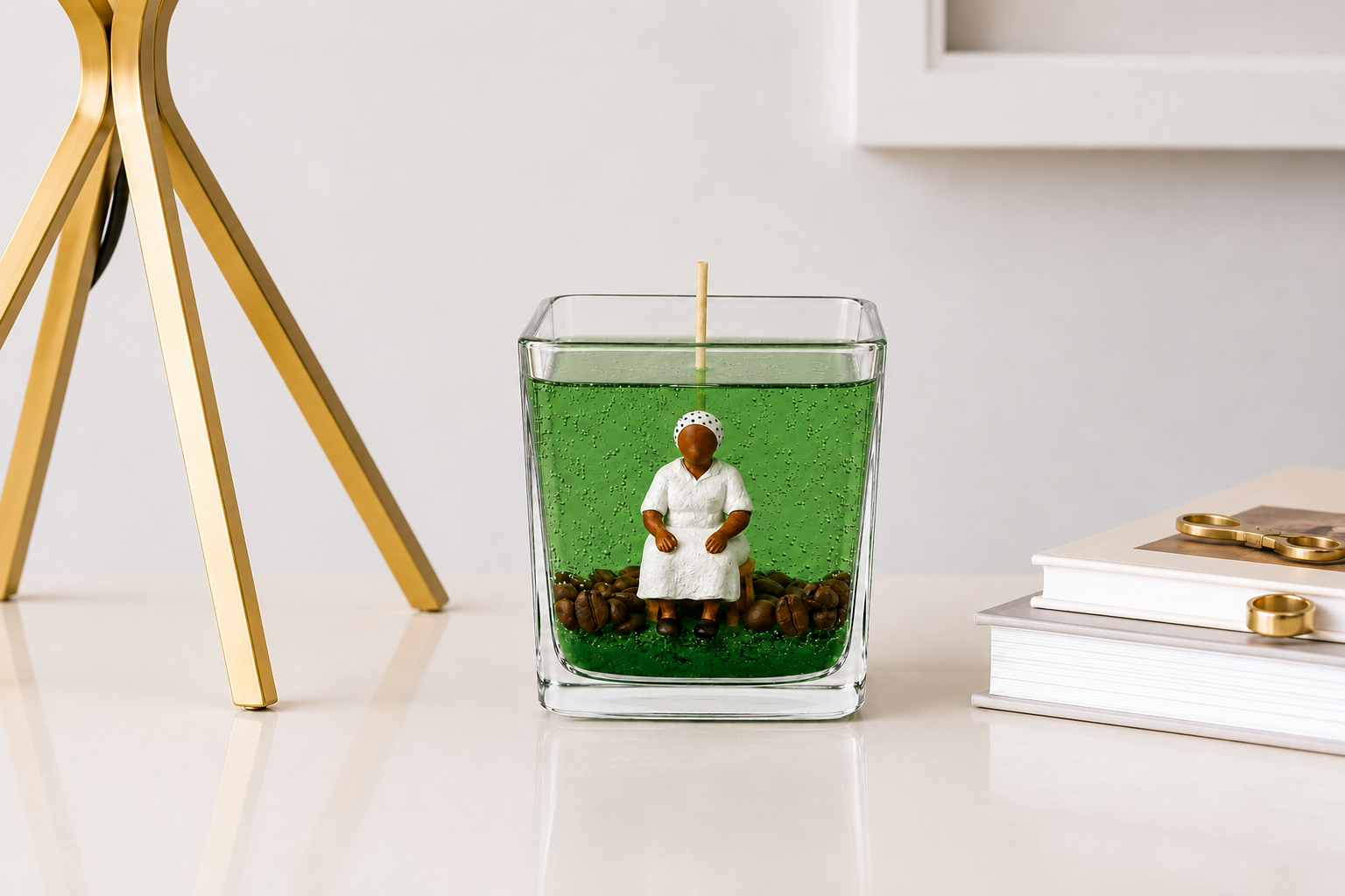 Vela em Gel Preta Velha artesanal em copo de vidro quadrado com parafina em gel verde translúcida, escultura de Preta Velha sentada e grãos de café decorativos. A peça está sobre uma mesa clara em ambiente sofisticado, acompanhada por livros e detalhes decorativos dourados.