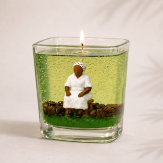 Vela em Gel Preta Velha artesanal em copo de vidro quadrado com parafina em gel verde translúcida, escultura de Preta Velha sentada e grãos de café decorativos. A vela está acesa e posicionada sobre fundo claro com iluminação suave e sombras delicadas de folhas.