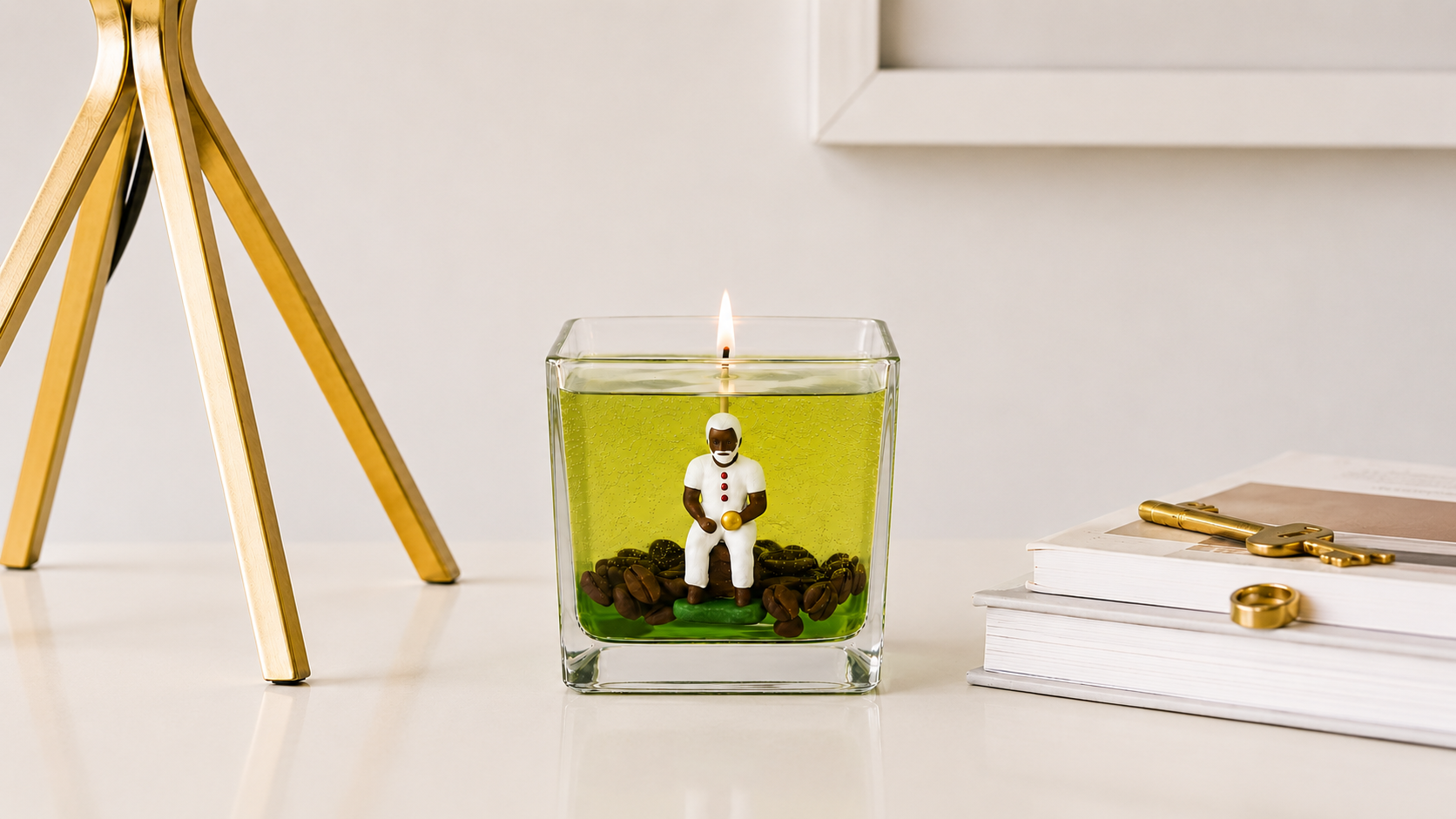 Vela em Gel Preto Velho artesanal em copo de vidro quadrado com parafina em gel verde translúcida, escultura de Preto Velho sentado, grãos de café decorativos e chama acesa. A peça está posicionada sobre uma mesa clara em ambiente sofisticado, com livros e detalhes decorativos dourados ao redor.