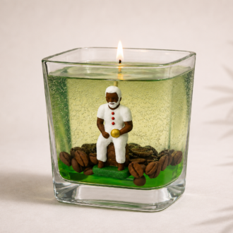 Vela em Gel Preto Velho artesanal em copo de vidro quadrado com parafina em gel verde translúcida, escultura de Preto Velho sentado, grãos de café decorativos e chama acesa. A peça está sobre fundo claro com iluminação suave e sombras delicadas de folhagens ao fundo.