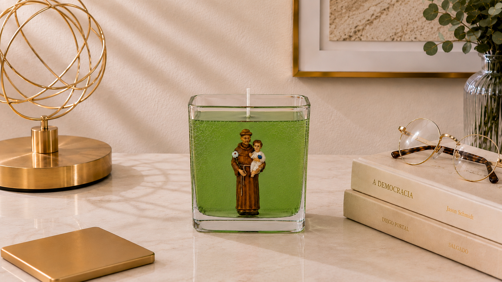 Vela em gel verde transparente com imagem de Santo Antônio segurando o Menino Jesus em recipiente quadrado de vidro. A peça está posicionada sobre uma superfície clara em ambiente decorado com luminária dourada, livros, óculos e elementos elegantes ao fundo, transmitindo uma atmosfera de fé, proteção e sofisticação.
