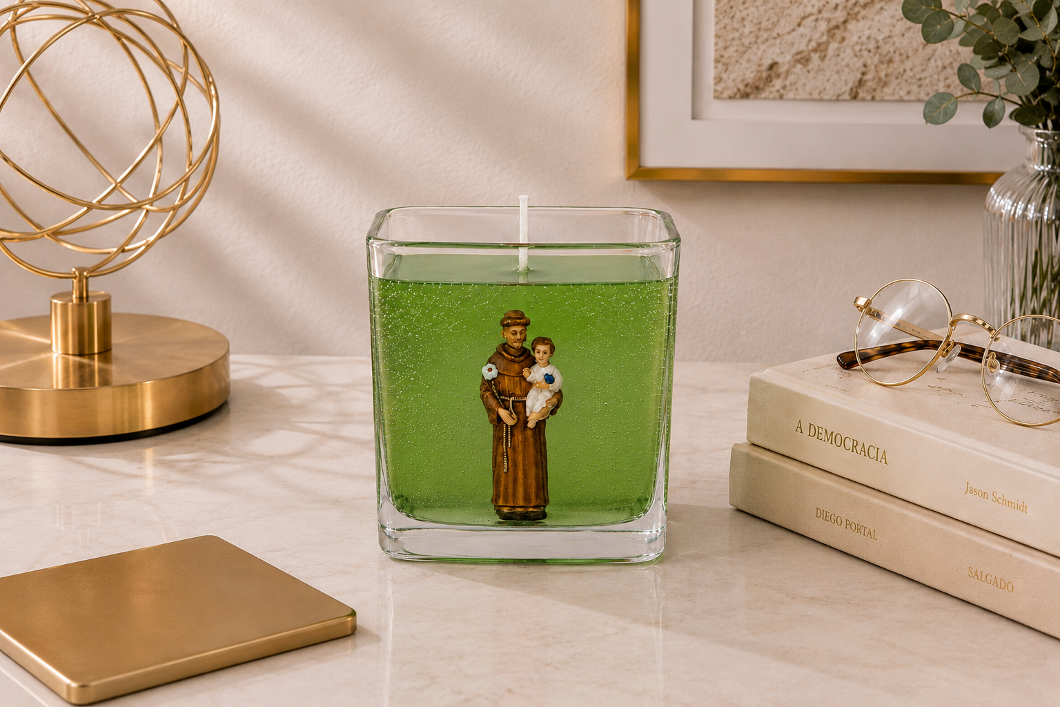 Vela em gel verde transparente com imagem de Santo Antônio segurando o Menino Jesus em recipiente quadrado de vidro. A peça está posicionada sobre uma superfície clara em ambiente decorado com luminária dourada, livros, óculos e elementos elegantes ao fundo, transmitindo uma atmosfera de fé, proteção e sofisticação.