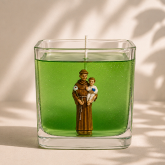 Vela em gel verde transparente com imagem de Santo Antônio segurando o Menino Jesus em recipiente quadrado de vidro. A peça está centralizada sobre fundo claro com iluminação suave e sombras delicadas de folhagens, transmitindo uma atmosfera de paz, devoção e elegância.