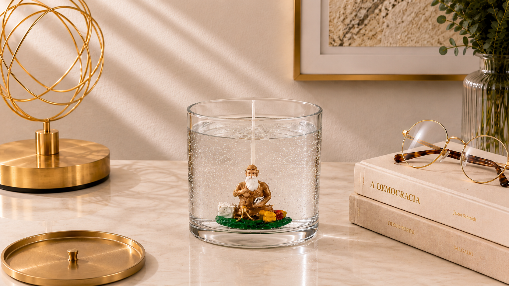 Vela em Gel Xangô artesanal em copo de vidro transparente, com representação decorativa de Xangô em seu interior, fotografada em ambiente sofisticado com livros, óculos, elementos dourados e iluminação natural suave.
