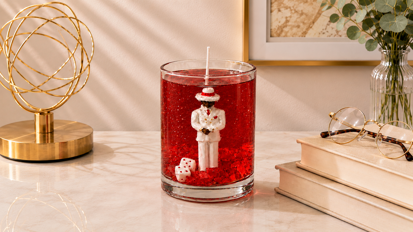 Vela em gel vermelho transparente com escultura de Zé Pilintra vestindo terno branco, gravata vermelha e chapéu característico. A peça está posicionada sobre uma mesa de mármore em ambiente sofisticado, acompanhada por livros decorativos, óculos e elementos dourados que reforçam sua elegância.