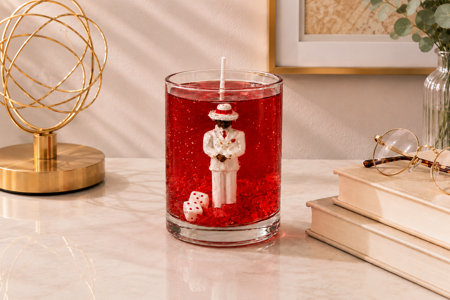 Vela em gel vermelho transparente com escultura de Zé Pilintra vestindo terno branco, gravata vermelha e chapéu característico. A peça está posicionada sobre uma mesa de mármore em ambiente sofisticado, acompanhada por livros decorativos, óculos e elementos dourados que reforçam sua elegância.