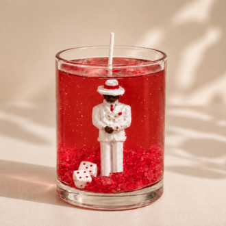 Vela em gel vermelho transparente com escultura de Zé Pilintra vestindo terno branco, gravata vermelha e chapéu característico. A composição inclui cristais vermelhos e dados decorativos na base do recipiente de vidro, destacando elegância, proteção e simbolismo espiritual.