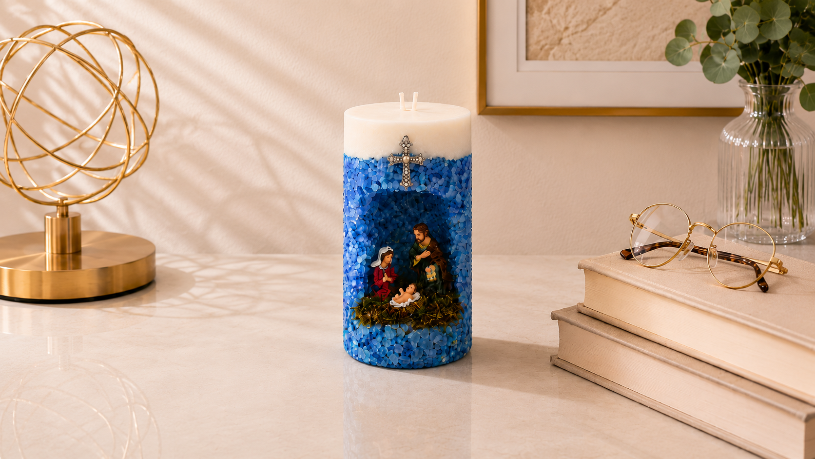 Vela em Gruta Sagrada Família artesanal decorada em tons de azul, com nicho central contendo as imagens de Maria, José e o Menino Jesus. A peça possui acabamento texturizado com pequenas pedras azuis, cruz metálica decorativa na parte frontal e dois pavios, apresentada em ambiente sofisticado com livros e elementos decorativos dourados.