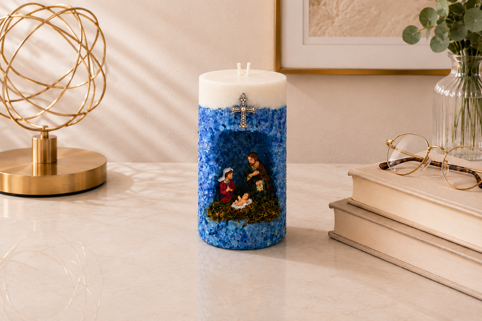 Vela em Gruta Sagrada Família artesanal decorada em tons de azul, com nicho central contendo as imagens de Maria, José e o Menino Jesus. A peça possui acabamento texturizado com pequenas pedras azuis, cruz metálica decorativa na parte frontal e dois pavios, apresentada em ambiente sofisticado com livros e elementos decorativos dourados.
