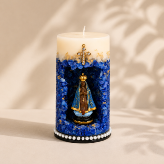 Vela Gruta de Nossa Senhora Aparecida artesanal em tons de azul intenso, branco e dourado, com imagem de Nossa Senhora Aparecida posicionada em nicho central decorativo. A peça apresenta cristais azuis, detalhes dourados aplicados manualmente, cruz ornamentada na parte superior e acabamento com pérolas na base. A vela está fotografada em ambiente minimalista de fundo claro, com iluminação suave e sombras naturais de folhagens.