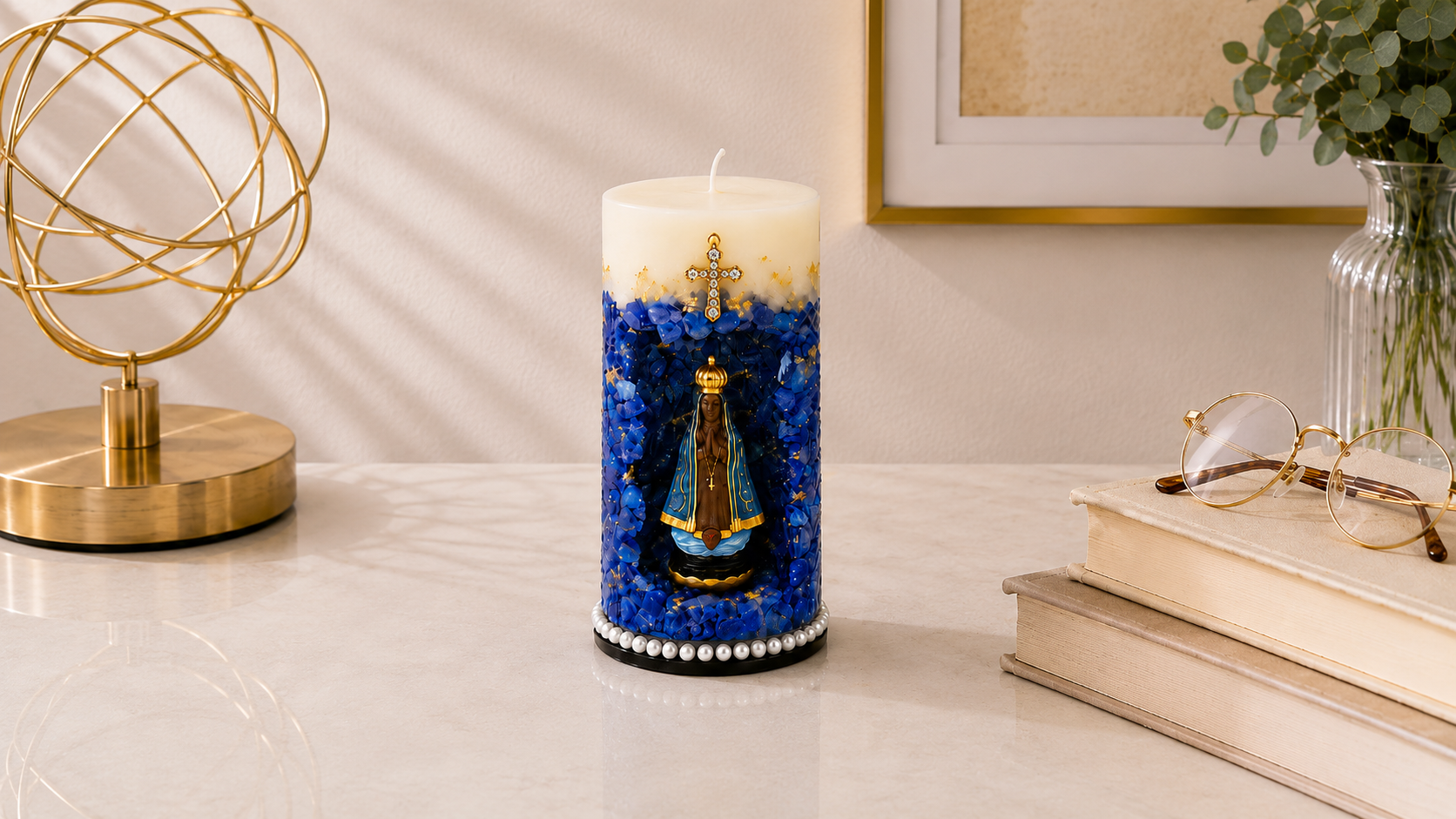 Vela Gruta de Nossa Senhora Aparecida artesanal em tons de azul profundo, branco e dourado, com imagem de Nossa Senhora Aparecida posicionada em um nicho central decorativo. A peça possui acabamento com cristais azuis, detalhes dourados, cruz ornamentada aplicada na parte superior e base adornada com pérolas. Está apresentada em ambiente sofisticado sobre mesa clara, acompanhada por livros, objeto decorativo dourado e vaso com folhagens.