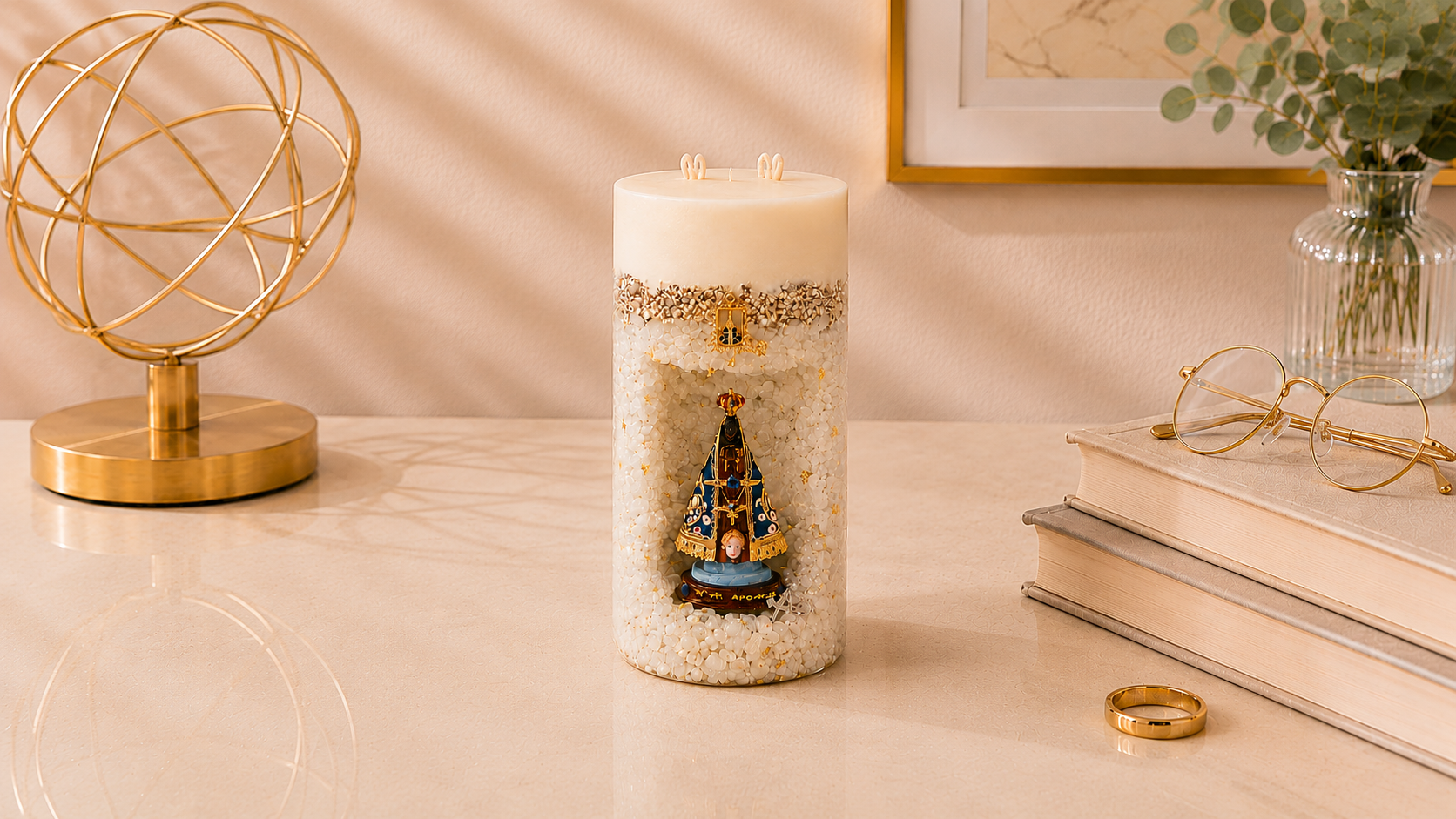 Vela Gruta de Nossa Senhora artesanal em formato cilíndrico, confeccionada com acabamento texturizado em tons claros e detalhes dourados. No centro da peça há um nicho decorativo com a imagem de Nossa Senhora Aparecida, destacada em azul e dourado. A vela está posicionada sobre uma superfície elegante em ambiente sofisticado, com livros, objeto decorativo dourado e elementos naturais ao fundo.