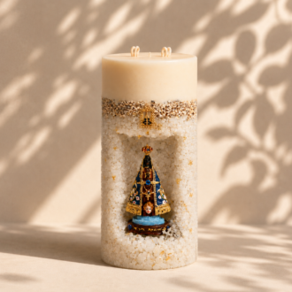 Vela Gruta de Nossa Senhora Aparecida artesanal em formato cilíndrico, confeccionada em tons claros com acabamento texturizado e delicados detalhes dourados. A peça possui um nicho central que abriga a imagem de Nossa Senhora Aparecida em azul, dourado e preto, destacando sua beleza e simbolismo religioso. A vela está posicionada sobre uma superfície clara, com iluminação suave e sombras naturais de folhagens ao fundo.