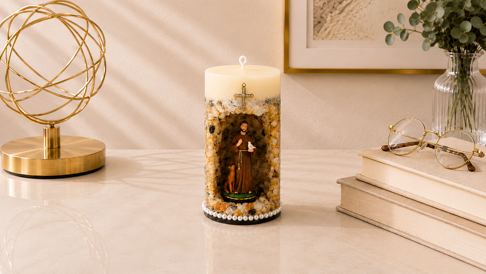 Vela Gruta de São Francisco de Assis artesanal em tons de bege, marfim e dourado, com imagem de São Francisco de Assis em nicho central decorativo acompanhado por um pequeno animal, símbolo de sua ligação com a natureza. A peça possui acabamento com pedras naturais em diferentes tonalidades terrosas, cruz ornamentada aplicada na parte superior e base decorada com pérolas. A fotografia apresenta a vela em ambiente elegante e acolhedor, com iluminação suave, livros decorativos e elementos dourados ao fundo.