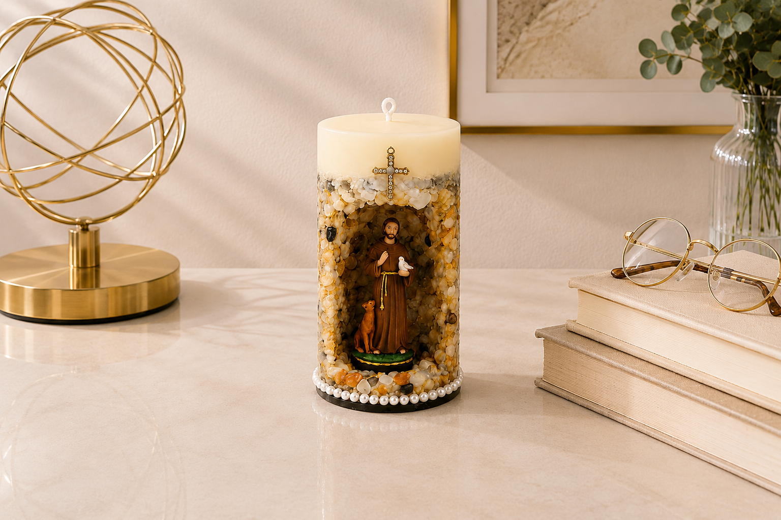 Vela Gruta de São Francisco de Assis artesanal em tons de bege, marfim e dourado, com imagem de São Francisco de Assis em nicho central decorativo acompanhado por um pequeno animal, símbolo de sua ligação com a natureza. A peça possui acabamento com pedras naturais em diferentes tonalidades terrosas, cruz ornamentada aplicada na parte superior e base decorada com pérolas. A fotografia apresenta a vela em ambiente elegante e acolhedor, com iluminação suave, livros decorativos e elementos dourados ao fundo.