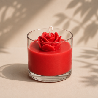 Vela Rosa Vermelha artesanal em copo de vidro transparente, com uma rosa vermelha esculpida em cera no topo. A imagem apresenta iluminação suave e natural, fundo bege claro com sombras delicadas de folhas, transmitindo romantismo, elegância e sofisticação.