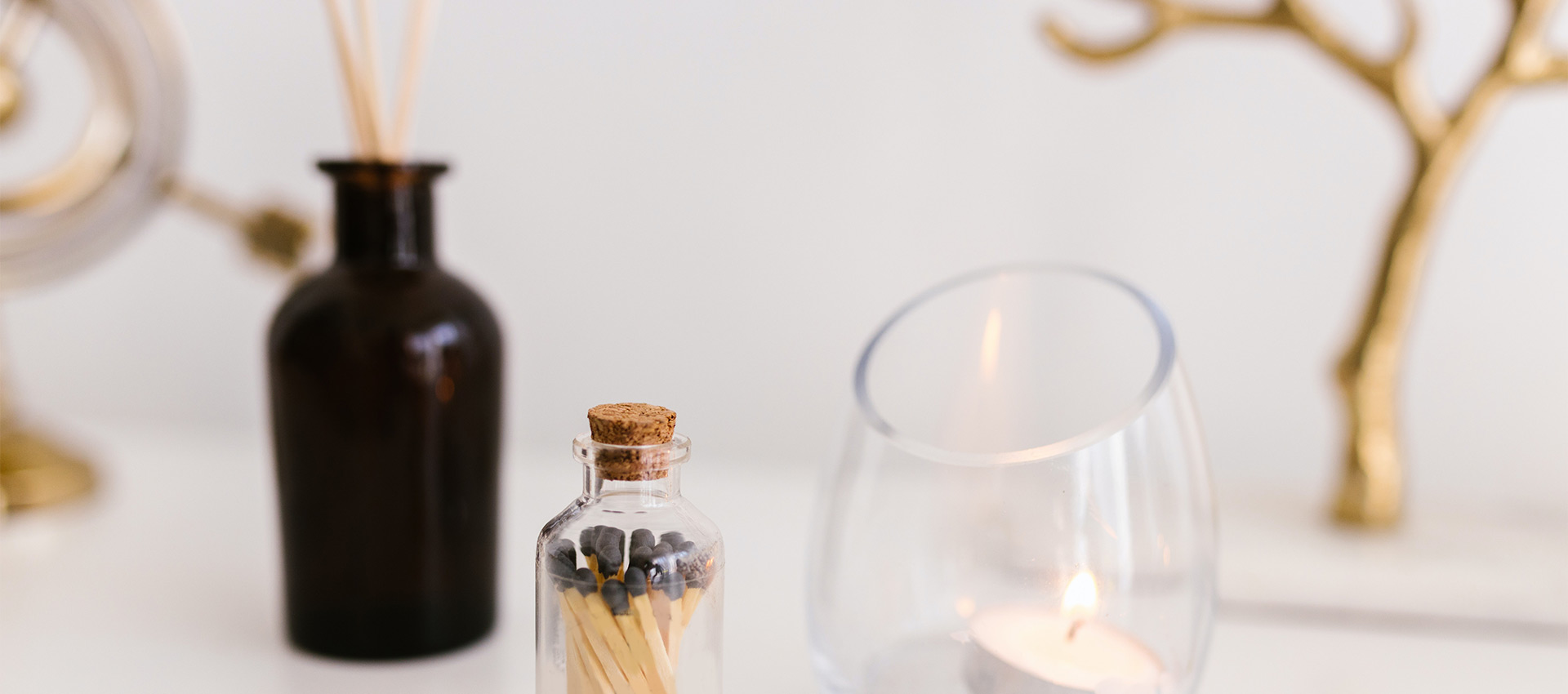 Composição elegante com vela acesa em recipiente de vidro, frasco decorativo com fósforos e difusor de aromas em vidro âmbar sobre superfície clara.